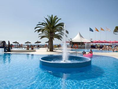 Pool mit Springbrunnen und Palmen vor dem Strand an einem sonnigen Tag.