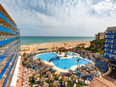 Vista de una piscina hotelera con tumbonas y playa al fondo bajo cielo nublado.