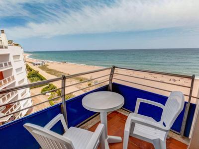 Balcón con sillas y mesa blancas, vista a la playa y el mar bajo un cielo despejado.