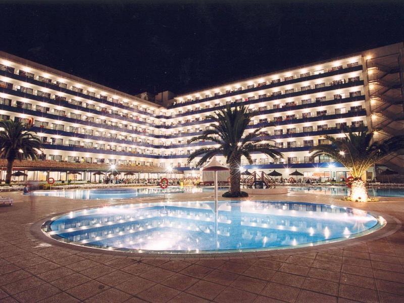 Hotel iluminado en forma de L con piscina circular y palmeras por la noche.