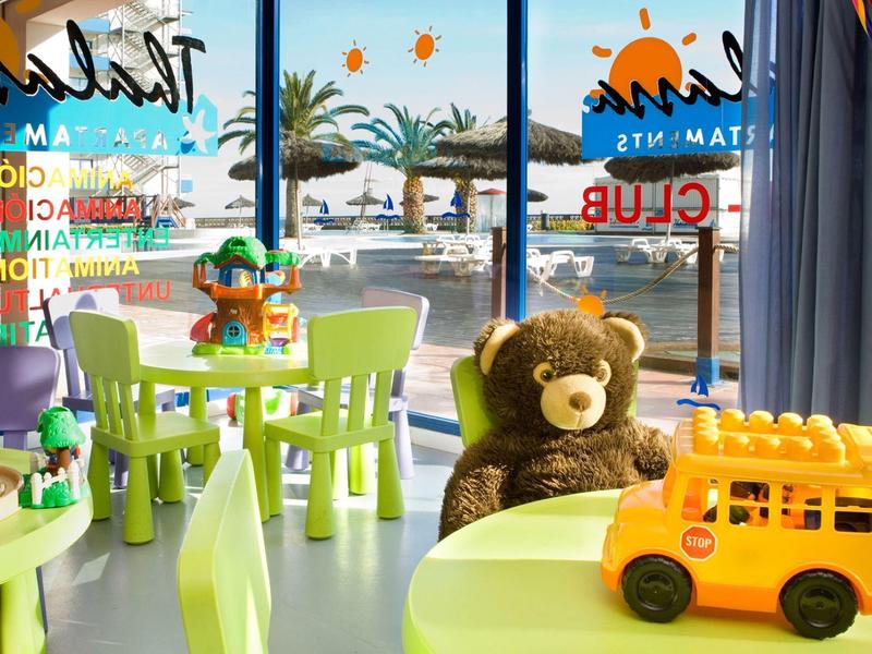 Vista de una sala de juegos colorida para niños con oso de peluche y juguetes junto a grandes ventanas al exterior.