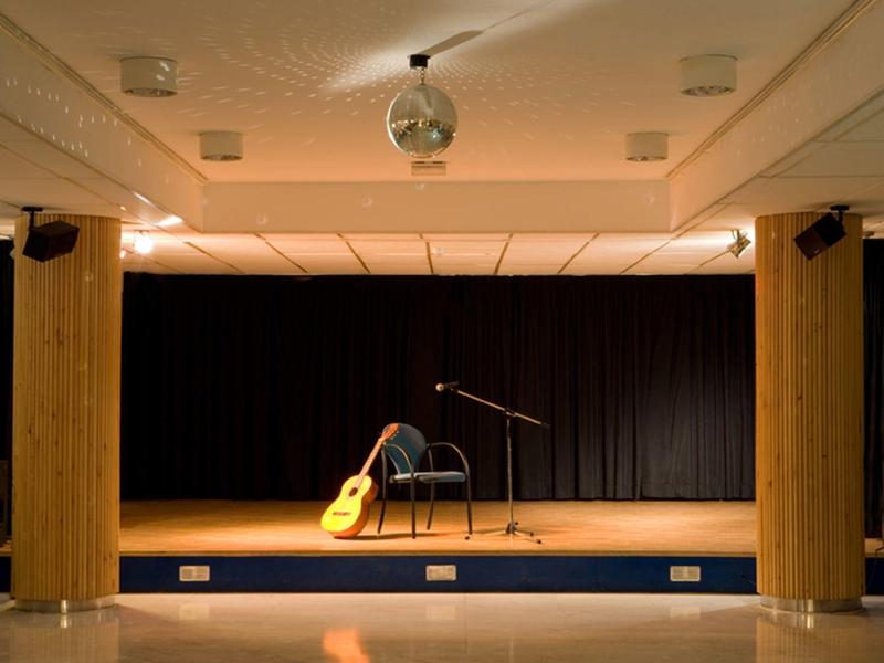 Escenario vacío con una silla, una guitarra y un micrófono frente a una cortina negra.