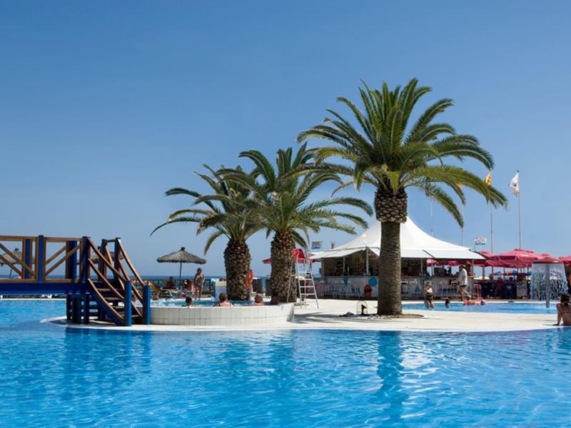 Una piscina con palmeras y un pequeño muelle bajo un cielo despejado, junto a sombrillas y asientos.