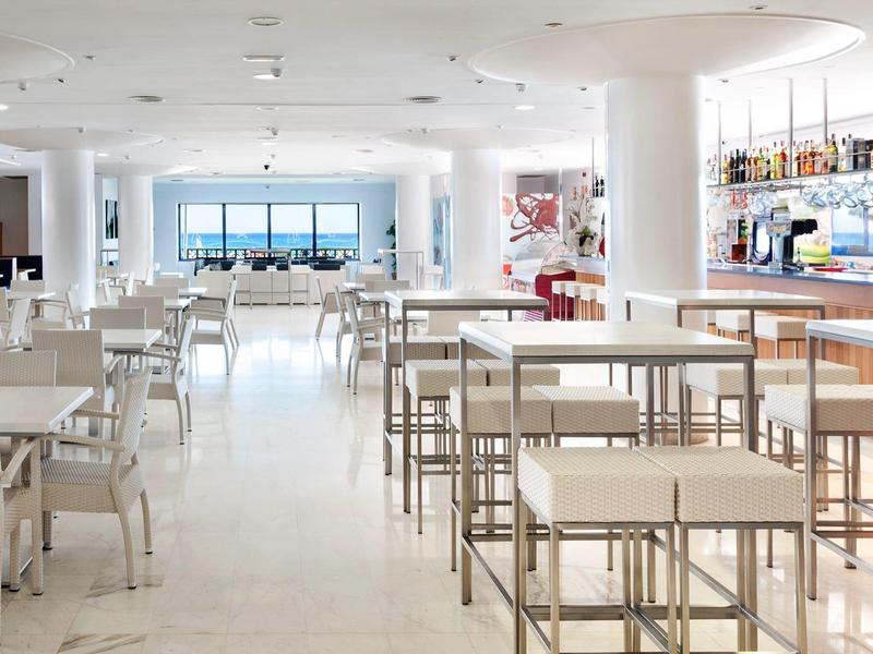 Comedor moderno y luminoso con mesas y sillas blancas y grandes ventanas con vista al mar.