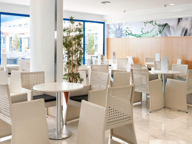 Restaurante moderno y luminoso en hotel con sillas blancas, mesas de madera y metal, y grandes ventanas.