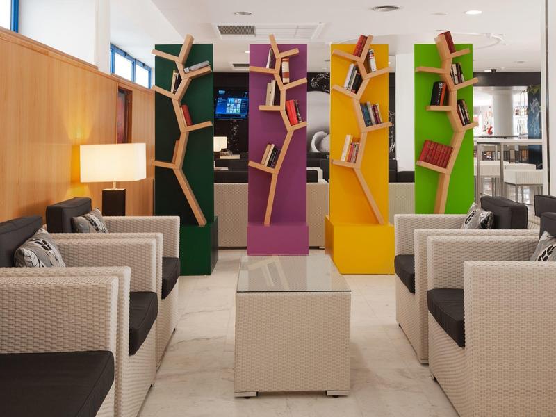 Moderna área de recepción con estanterías de colores, zonas de descanso y suelo blanco en un hotel.