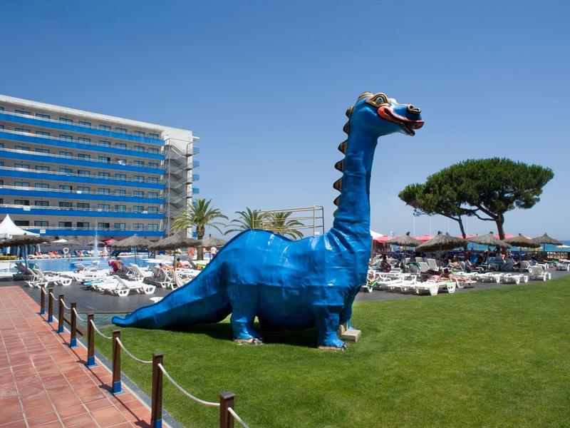 Figura grande de dinosaurio azul en el césped del hotel junto a tumbonas y un edificio hotelero.