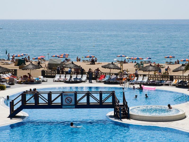 Vista de una playa con tumbonas, sombrillas y una piscina con un pequeño puente.