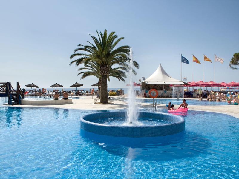 Una piscina azul con fuente, rodeada de tumbonas, palmeras y sombrillas en la playa.
