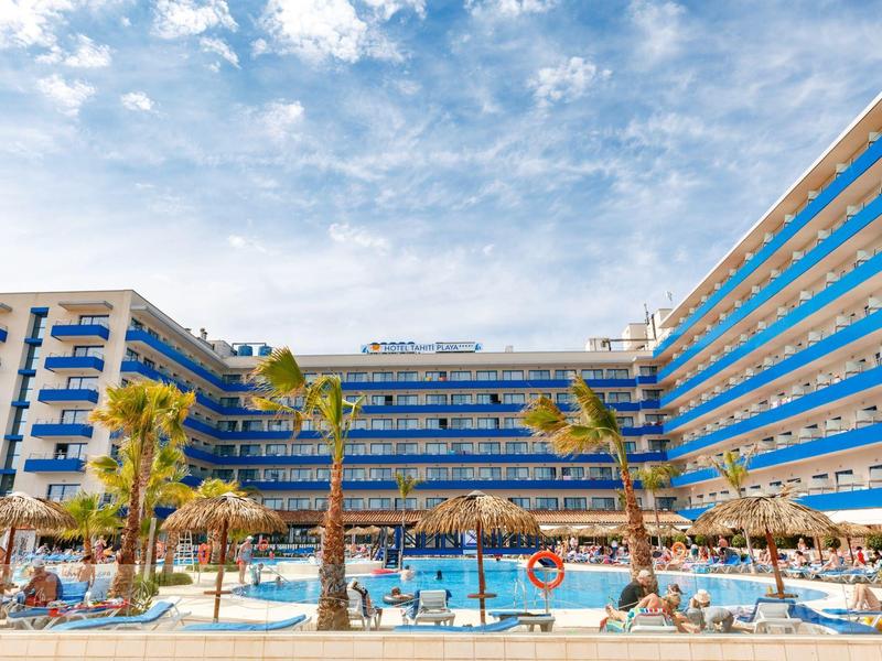 Gran hotel con piscina, palmeras y sol, rodeado de balcones azules y cielo claro.