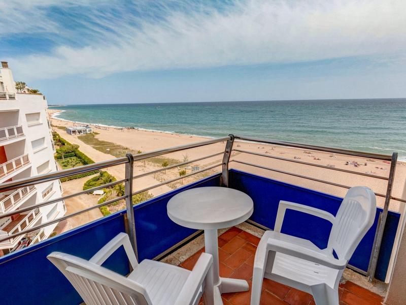 Balcón con sillas y mesa blancas, vista a la playa y el mar bajo un cielo despejado.