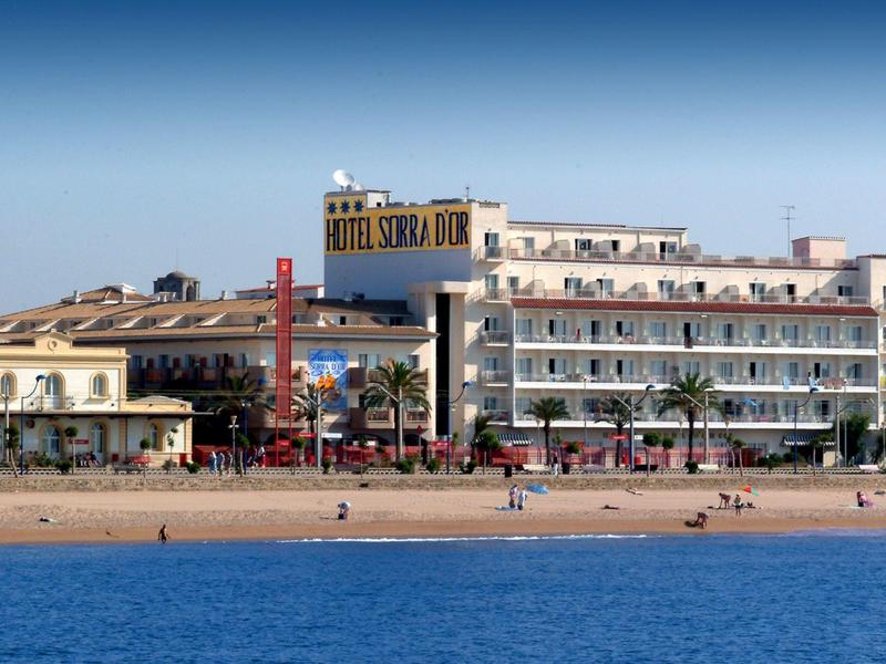Hotel de varios pisos en una playa con el mar en primer plano y cielo azul.