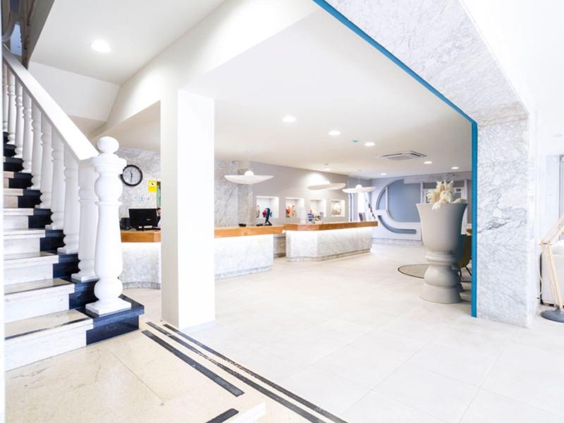 Área de entrada luminosa del hotel con recepción, escalera y decoración moderna.