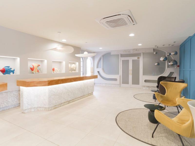 Recepción moderna con mostrador blanco, paredes claras y sillas amarillas junto a una pared azul.