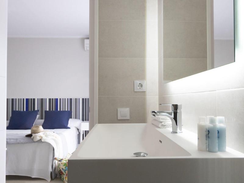 Baño moderno con lavabo blanco, espejo y vista a dos camas con cojines azules.