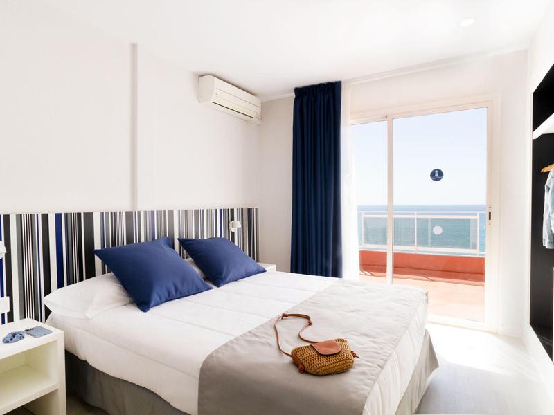 Dormitorio luminoso con cama doble, almohadas azules y acceso a balcón con vista al mar.