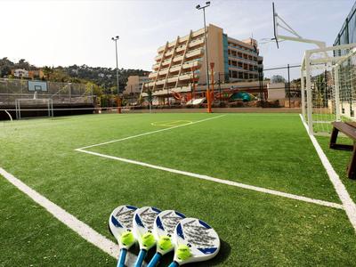 Fußball auf Kunstrasenplatz mit Gebäude und Hügel im Hintergrund.