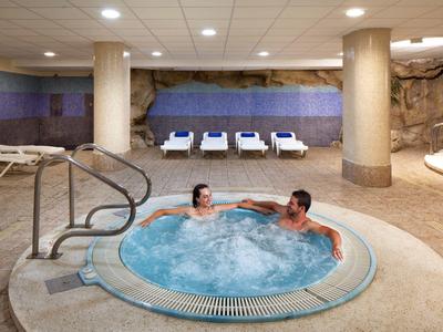 Zwei Personen entspannen im Whirlpool eines Spa mit Liegestühlen und Felswand im Hintergrund.