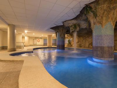 Innenbereich mit blau beleuchtetem Schwimmbad und Felsenwand an der Decke und Säulen.