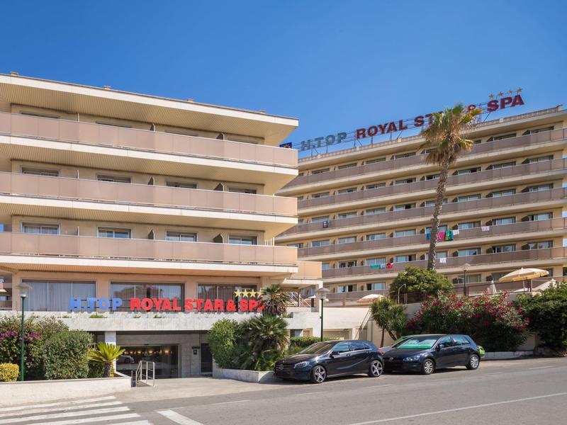 Hotel moderno con più piani e ampi balconi, auto parcheggiate e palme davanti.