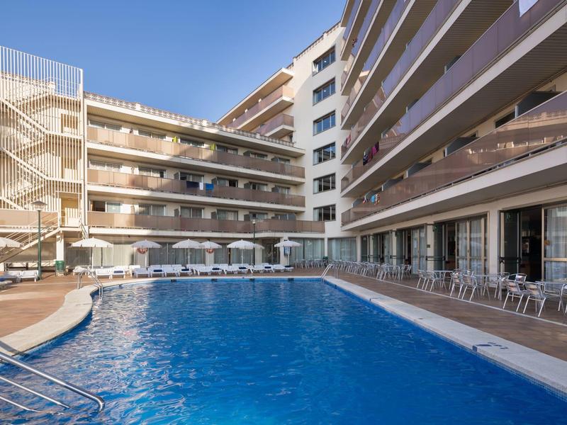 Area piscina moderna dell'hotel con lettini e ombrelloni, circondata da edifici su più piani.