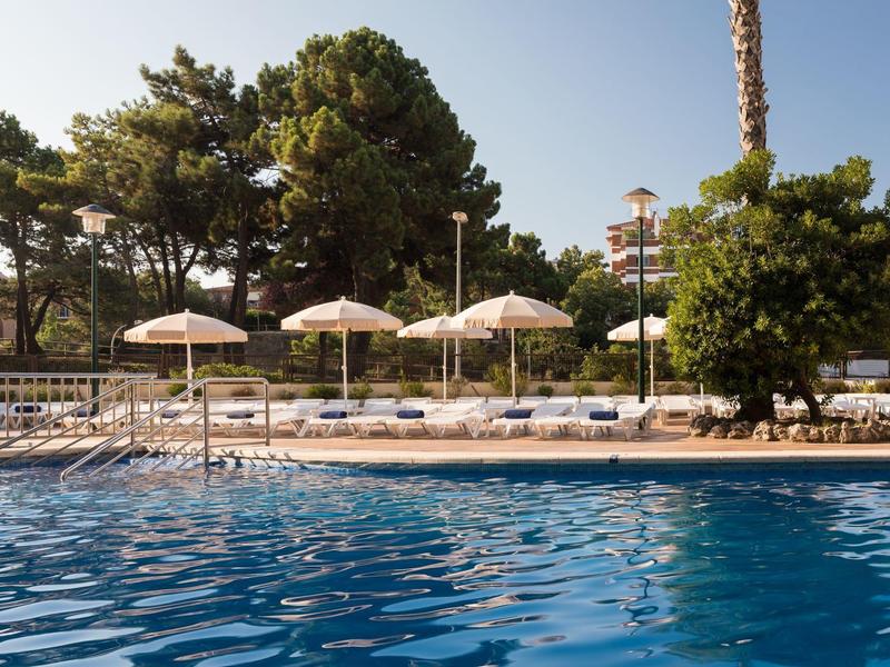 Piscina con acqua blu circondata da lettini e ombrelloni in un giardino d'hotel.