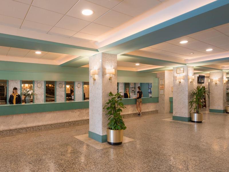 Ampia hall dell'hotel con reception, colonne multiple e piante sotto luci a soffitto luminose.