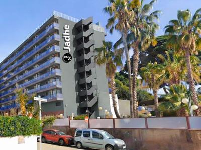 Mehrstöckiges Hotelgebäude mit Logo, umgeben von Palmen und geparkten Autos im Vordergrund.