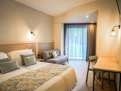 Helles Hotelzimmer mit Doppelbett, Sofa, Schreibtisch und Balkon mit Vorhängen.