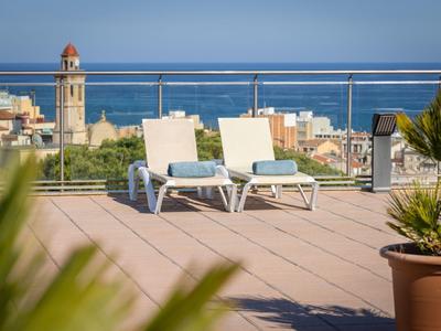 Zwei weiße Liegestühle auf einer sonnigen Terrasse mit Meerblick und Stadt im Hintergrund.