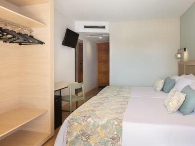 Hotelzimmer mit Doppelbett, offenem Kleiderschrank, Fernseher und schlichtem Dekor.