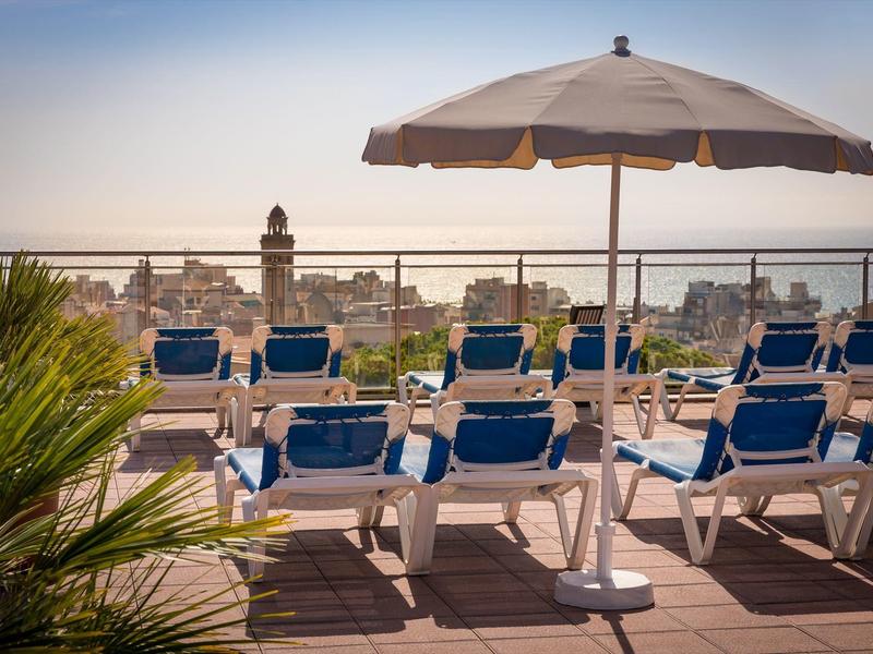 Střešní terasa s lehátky a slunečníkem s výhledem na město při západu slunce.