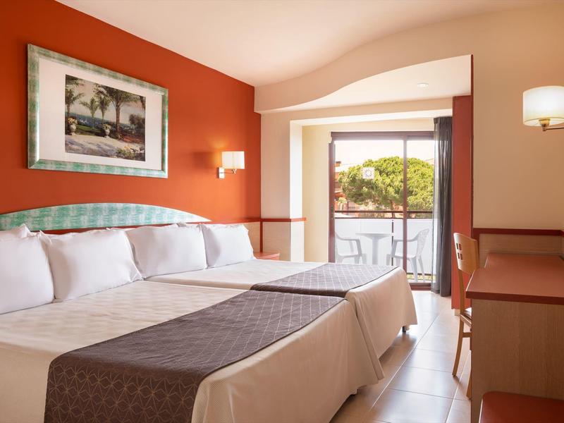 Světý pokoj hotelu s manželskou postelí, oranžovou zdí a balkonem s výhledem na stromy.