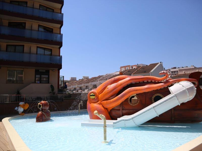 Dětské hřiště s vodní skluzavkou ve tvaru chobotnice vedle hotelové budovy pod modrou oblohou.