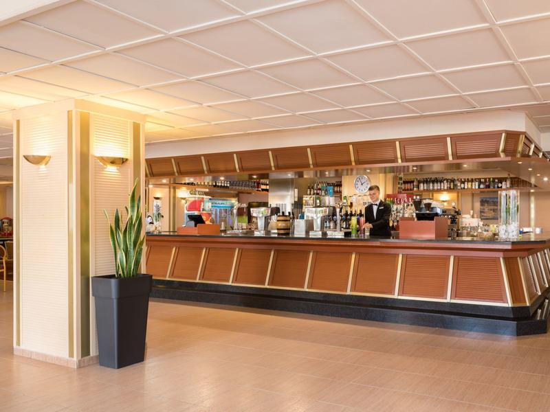 Elegantní barový prostor hotelu s barem, barmany a velkou rostlinou v popředí.