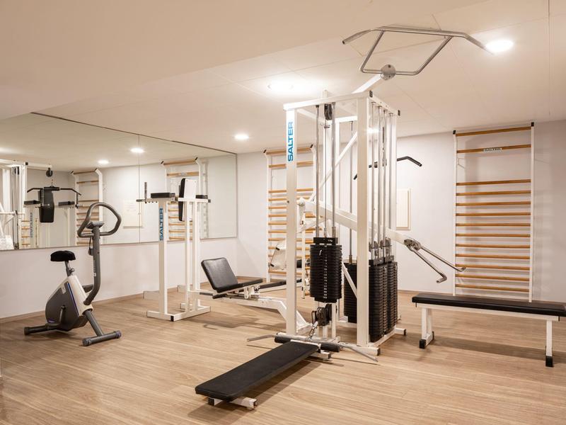 Světlá posilovna s různými tréninkovými zařízeními a dřevěnou podlahou.