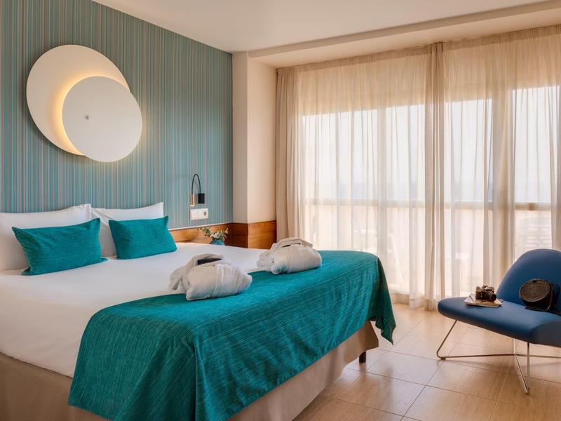 Modernes Hotelzimmer mit großem Bett, türkisfarbenem Überwurf und Balkon mit Vorhängen.