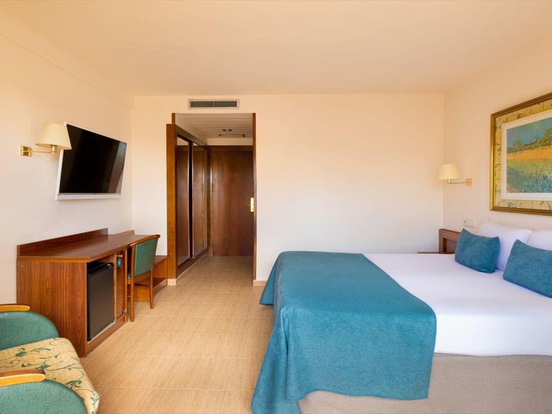 Ein helles Hotelzimmer mit Doppelbett, blauen Decken und Kissen, Fernseher und Schreibtisch.