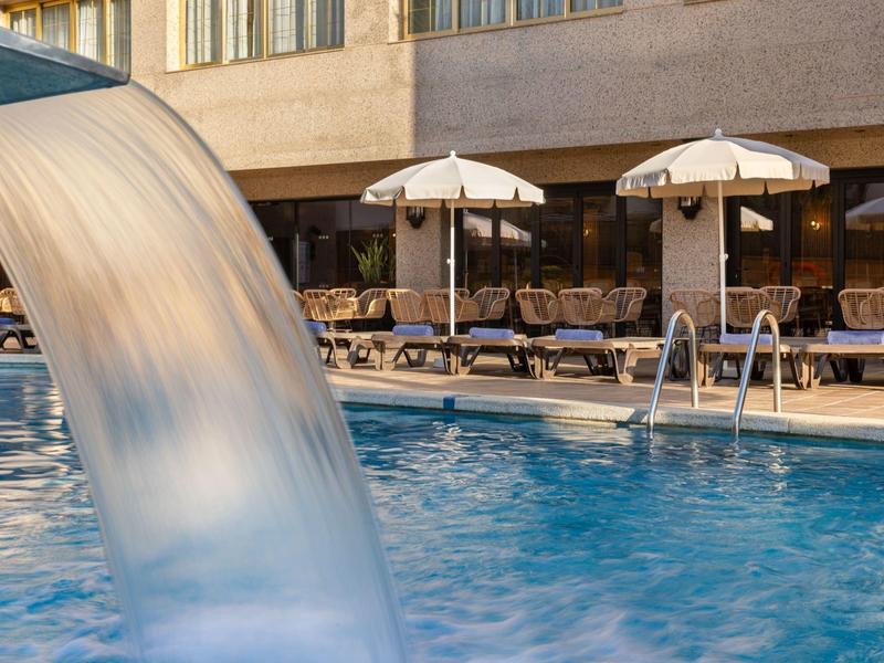Ein Wasserfall fließt in einen Pool mit Liegestühlen und Sonnenschirmen im Hintergrund.