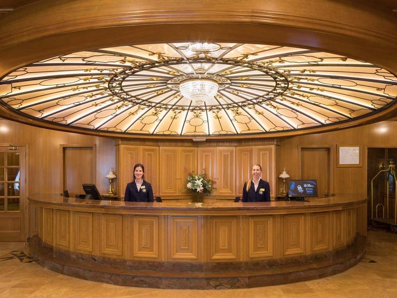 Hotelempfang mit rundem hölzernem Tresen und Rezeptionisten unter einer großen, dekorativen Deckenleuchte.