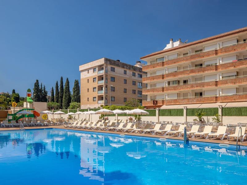 Grande piscina esterna con lettini davanti a un edificio alberghiero di più piani in una giornata di sole.