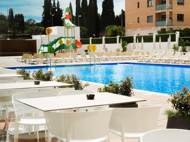 Area piscina esterna moderna con tavoli bianchi, sedie e scivolo accanto a un hotel.