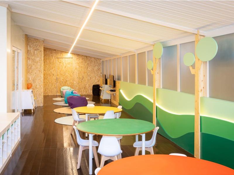 Area giochi e pranzo luminosa con tavoli colorati, sedie bianche e decorazioni murali.
