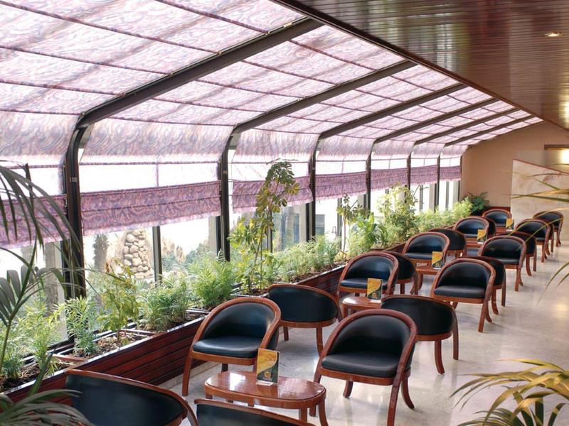 Overdekt zitgedeelte met rijen stoelen en tafels naast grote ramen en planten.