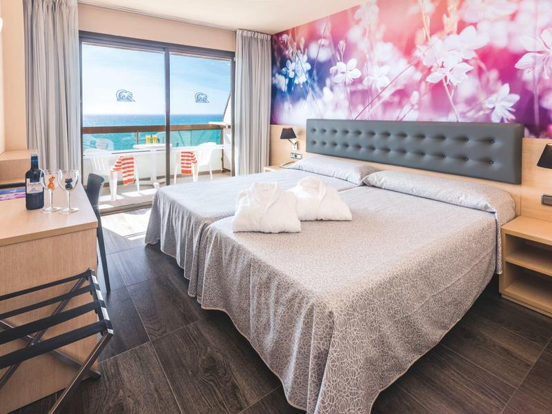 Lichte hotelkamer met tweepersoonsbed, bloemenmuurdecoratie en uitzicht op balkon en zee.