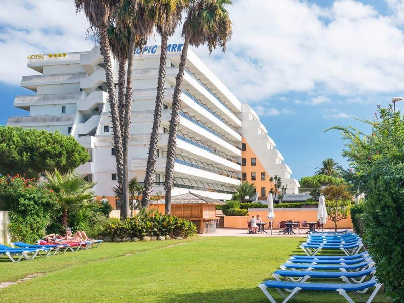 Modern hotel met palmbomen en ligstoelen op verzorgd grasveld onder blauwe lucht.