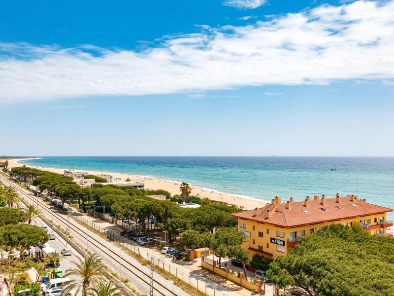 Uitzicht op een strand met spoorlijnen, bomen en een geel gebouw onder een blauwe lucht.