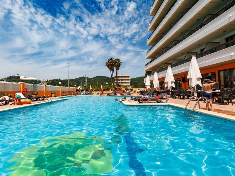 Groot buitenzwembad met ligstoelen en parasols naast een hotel op een zonnige dag.