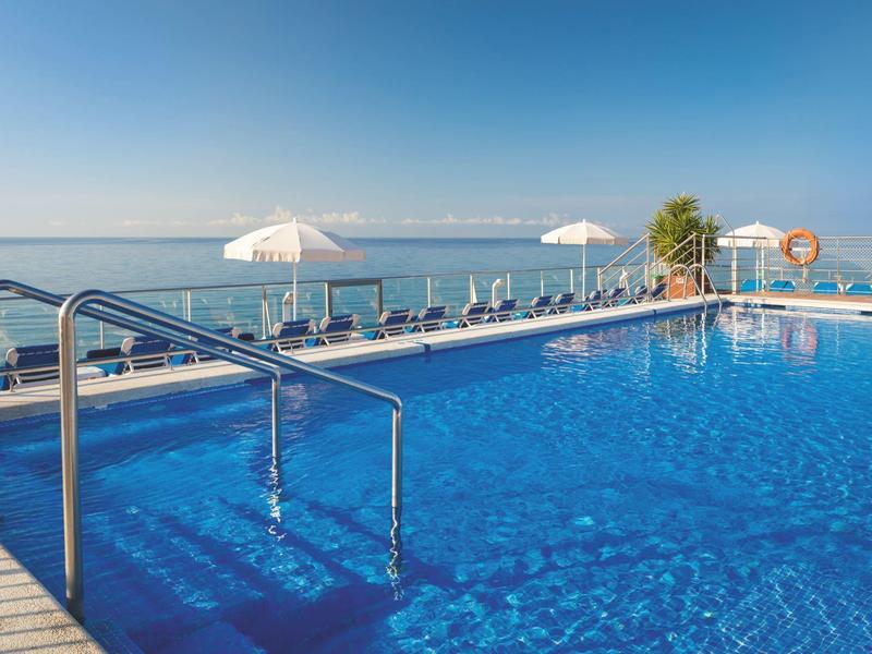 Gran piscina con agua azul clara, tumbonas y sombrillas con vistas al mar.