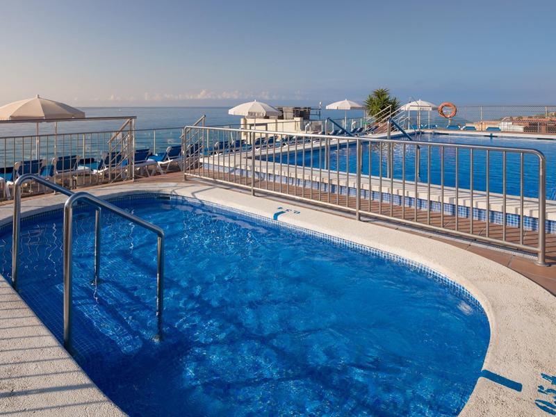 Dos piscinas con vista al mar, rodeadas de sombrillas y una barandilla en el hotel.